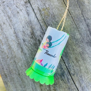 May include: A white cylinder ornament with a green scalloped edge. It features a colorful illustration of two children on a swing with the word "Pomander" printed on the side.