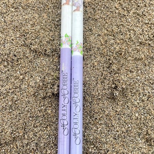 Peut inclure: Deux crayons vintage Holly Hobbie avec des embouts verts et rouges. Les crayons ont un corps blanc avec un motif floral et un dessin d'une fille. Les crayons sont violets en bas et reposent sur du sable.