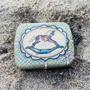 May include: A rectangular metal tin featuring a central oval illustration of a rocking horse with a red saddle. The background of the tin has a yellow and green woven pattern, accented by a pink ribbon design on the right side and a scalloped border.