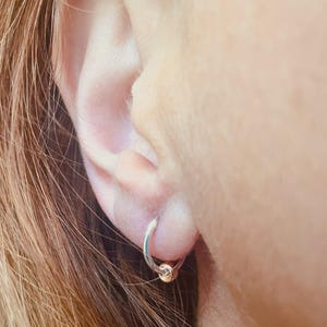 May include: A close-up of a silver hoop earring with a small gold bead. The earring is in a person's ear, with the ear and hair visible in the frame. The earring is simple and elegant.