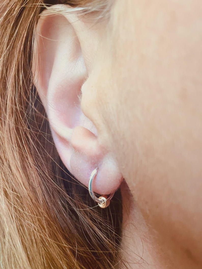 May include: A close-up of a silver hoop earring with a small gold bead. The earring is in a person's ear, with the ear and hair visible in the frame. The earring is simple and elegant.