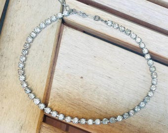 May include: A silver-toned necklace with a row of clear, sparkling rhinestones. The necklace is laid out on a wooden surface, showcasing its delicate design and reflective qualities. The clasp is visible.