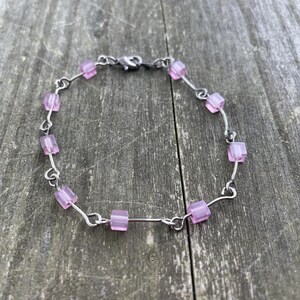 Könnte beinhalten: Ein zartes Armband mit kleinen, hellvioletten quadratischen Perlen, die auf einem silberfarbenen Draht aufgereiht sind. Das Armband hat ein schlichtes Design mit einem Verschluss. Die Perlen sind gleichmäßig verteilt.