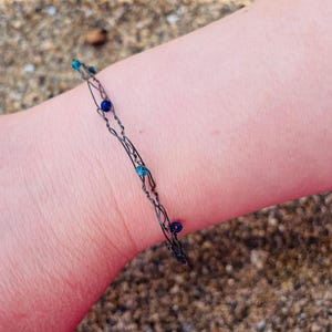 Puede incluir: Una pulsera delicada construida con alambre metálico oscuro y fino, trenzado en un diseño de doble hebra. Pequeñas cuentas redondas en varios tonos de azul y verde azulado están intercaladas a lo largo del alambre, aseguradas en la estructura.