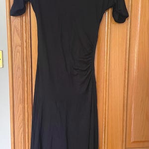 May include: A solid black midi-length dress featuring a high mock turtleneck collar, short sleeves, and a gathered side detail at the waist that creates soft draping across the torso. The skirt falls to a flared hemline with a smooth finish.