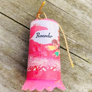 May include: A pink and white pomander ornament with a gold string. The ornament has a cartoon image of a person with a yellow shirt and a green skirt. The ornament says "Pomander" on the front.