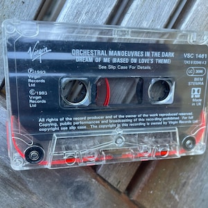 May include: A clear plastic cassette tape featuring a black label with white text reading Orchestral Manoeuvres in the Dark, Dream of Me, Based on Love's Theme, Virgin Records, 1993, VSC 1461, and Made in UK, with visible red internal tape reels.