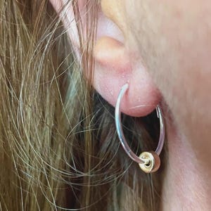 May include: A close-up of a silver hoop earring with a small gold bead. The earring is worn in an ear, with the person's hair visible in the background. The earring is simple and elegant.
