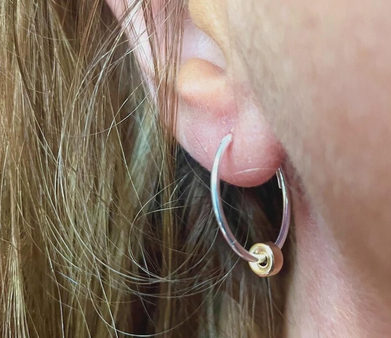 May include: A close-up of a silver hoop earring with a small gold bead. The earring is worn in an ear, with the person's hair visible in the background. The earring is simple and elegant.
