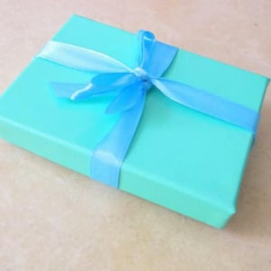 May include: A rectangular, turquoise gift box tied with a light blue satin ribbon and bow. The box is closed and sits on a light beige surface. The ribbon is wrapped around the box and tied in a bow on top.