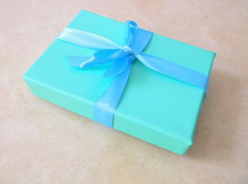 May include: A rectangular, turquoise gift box tied with a light blue satin ribbon and bow. The box is closed and sits on a light beige surface. The ribbon is wrapped around the box and tied in a bow on top.