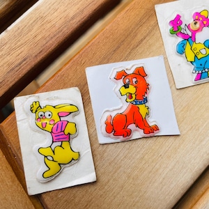 May include: Three vintage stickers featuring cartoon characters. One sticker shows a yellow rabbit in a pink shirt, another depicts an orange dog with a blue collar, and the third shows a blue and pink character holding a flower.