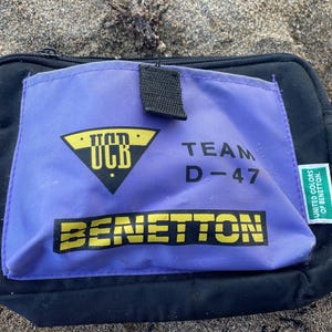Puede incluir: Bolso rectangular negro con un bolsillo morado con el texto amarillo "BENETTON". El bolsillo también muestra "UCB" en un triángulo amarillo y "TEAM D-47". Una etiqueta verde dice "UNITED COLORS OF BENETTON".