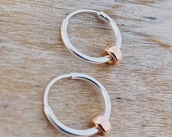 May include: Two silver hoop earrings with a small, rose gold bead accent. The earrings are displayed on a light brown wooden surface. The hoops are round and have a polished finish, with a simple, elegant design.