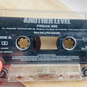 May include: A clear cassette tape with a black label featuring the text "ANOTHER LEVEL" and "FREAK ME." The label includes track listings and the text "SIDE A." The cassette is held by a person.