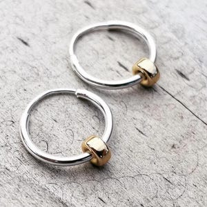 Op de afbeelding: Twee zilveren oorringen met een gouden kraal accent. De oorbellen zijn cirkelvormig en hebben een eenvoudig, elegant ontwerp. De gouden kraal voegt een vleugje contrast en verfijning toe aan de oorbellen.