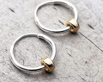 May include: Two silver hoop earrings with a gold bead accent. The earrings are circular and have a simple, elegant design. The gold bead adds a touch of contrast and sophistication to the earrings.