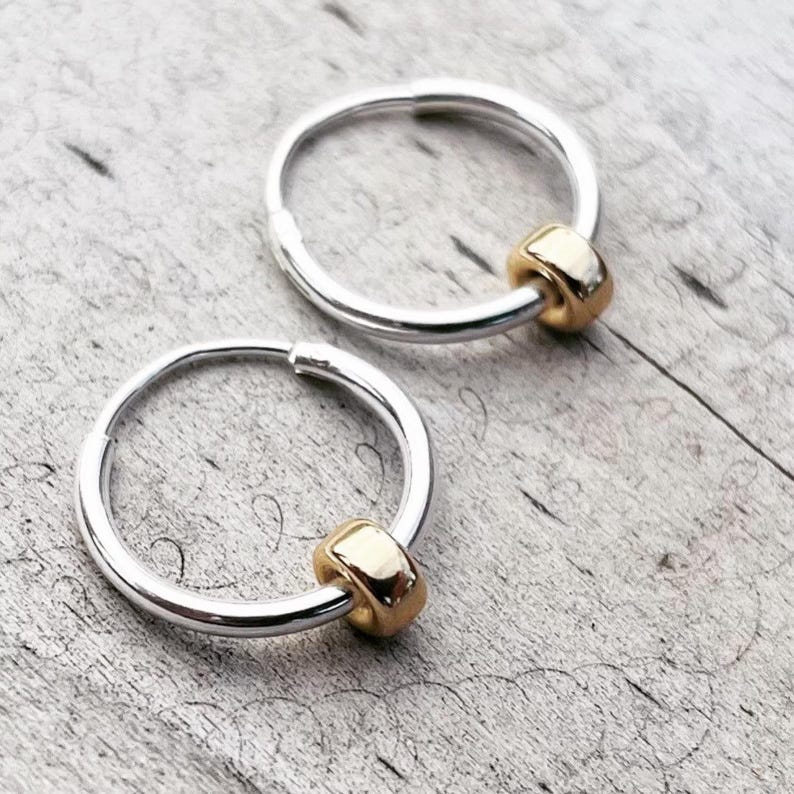 May include: Two silver hoop earrings with a gold bead accent. The earrings are circular and have a simple, elegant design. The gold bead adds a touch of contrast and sophistication to the earrings.
