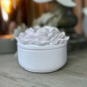 May include: A small, round, white ceramic trinket box with a detailed, three-dimensional rose design on the lid. The box is smooth and matte, with a slightly rounded base. The rose petals are intricately sculpted, adding a delicate touch.