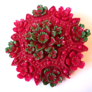 May include: A vintage, red brooch with a floral design. The brooch features a central cluster of green and red flowers surrounded by a detailed pattern of smaller flowers and raised dots. The brooch has a textured surface and a slightly raised, dimensional appearance.