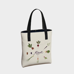 Könnte beinhalten: Eine weiße Canvas-Tragetasche mit schwarzen Henkeln und einem Design aus verschiedenen Wurzelgemüsen in einem Kreis um das Wort "Root" in lila Schrift.