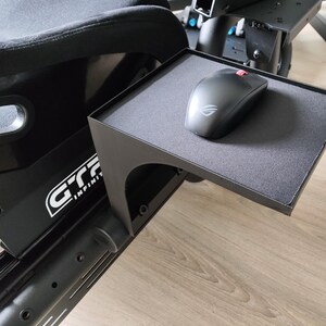 Puede incluir: Un ratón de ordenador negro con un acento rojo descansa sobre una bandeja rectangular negra sujeta a una silla negra de simulador de carreras. La bandeja tiene una superficie texturizada. La silla tiene el texto "GTR INFINITY" en blanco.