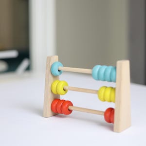 May include: Wooden abacus with three rows of colourful beads. The top row has blue beads, the middle row has yellow beads, and the bottom row has red beads. The frame is made of natural wood.