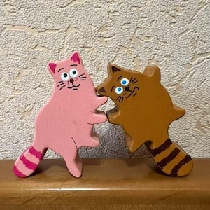 May include: Two wooden cat figurines. One is pink with a striped tail, the other is brown with a striped tail. Both have smiling faces and are touching noses. The figurines are on a wooden surface.