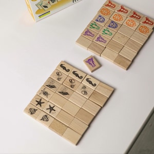 Può includere: Gioco di abbinamento in legno con illustrazioni di animali e creature marine. Il gioco presenta piccoli blocchi di legno quadrati con disegni colorati di leoni, giraffe, cavallucci marini e altro. I blocchi sono disposti su una superficie bianca.