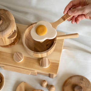 May include: Wooden toy kitchen set featuring a frying pan with a wooden egg, a pot, and other accessories. The set is made of light-colored wood and includes a wooden cutting board and small wooden circles.