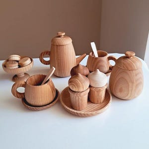 Puede incluir: Un juego de té de madera con tetera, tazas, platillos y un cuenco. El juego incluye un azucarero, una cuchara y pasteles de madera en miniatura. Los artículos son de madera clara y están dispuestos sobre una superficie blanca.
