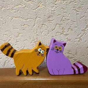 May include: Two wooden cat figurines. One is orange with a striped tail, the other is purple with a striped tail. Both have painted faces with whiskers and large eyes. They are standing on a wooden surface.