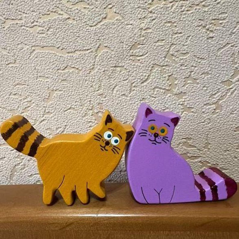 May include: Two wooden cat figurines. One is orange with a striped tail, the other is purple with a striped tail. Both have painted faces with whiskers and large eyes. They are standing on a wooden surface.