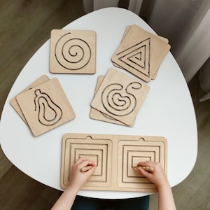 May include: Wooden tracing boards with various shapes, including a spiral, triangle, and a pear. The boards are light brown and designed for children's fine motor skill development. A child's hands are shown using a wooden stylus on a square tracing board.