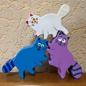 May include: Three wooden cat figurines in white, blue, and purple. The white cat has a striped tail and yellow eyes. The blue and purple cats have round eyes and striped tails. The cats are arranged in a playful, overlapping design.