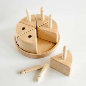 Könnte beinhalten: Holzspielzeugkuchen mit sechs herausnehmbaren Stücken und Holzkerzenstiften. Der Kuchen steht auf einem runden Holztablett. Ein Stück ist vom Kuchen getrennt.