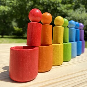 Puede incluir: Un juego de juguetes de madera de colores del arcoíris con figuras cilíndricas rematadas con esferas. El juego incluye piezas rojas, naranjas, amarillas, verdes, azules y moradas. Las piezas están dispuestas sobre una superficie de madera clara.