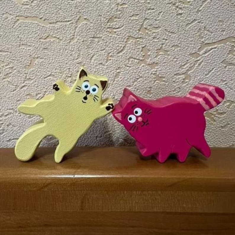 May include: Two wooden cat figurines. One is yellow with black details, outstretched paws, and a surprised expression. The other is magenta with a striped tail and a curious expression. Both are on a wooden surface.