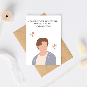 Pu&ograve; includere: Un biglietto di auguri bianco con il testo "I couldn't get you Conrad, so I got you this card instead." Il biglietto presenta un'illustrazione di una persona e piccoli cuori rosa. Il biglietto &egrave; su una busta marrone.