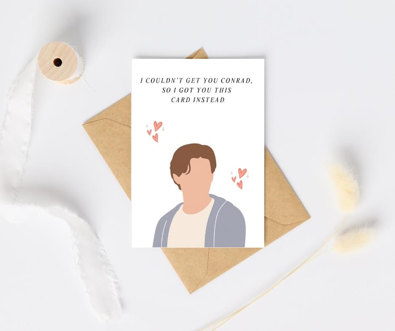 Pu&ograve; includere: Un biglietto di auguri bianco con il testo "I couldn't get you Conrad, so I got you this card instead." Il biglietto presenta un'illustrazione di una persona e piccoli cuori rosa. Il biglietto &egrave; su una busta marrone.