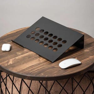 May include: A black metal laptop stand with a series of circular cutouts. The stand is on a round, dark wood table, with a white wireless mouse and a white wireless earbud case nearby.