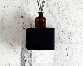 May include: A black toilet paper holder with a roll of white toilet paper. Above the holder is a brown glass bottle with reed diffuser sticks. The background is a white wall with gray marbling.