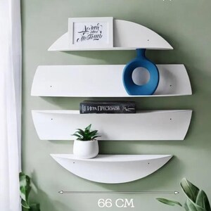 May include: White, semi-circular wall shelves with a total width of 66 cm. The shelves hold a framed picture, a blue vase, a book, and a small potted plant. The shelves are mounted on a light green wall.