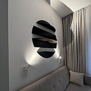 May include: A modern wall-mounted light fixture with a unique design. The fixture is composed of several black, curved, layered elements, creating a three-dimensional effect. The fixture is mounted on a white wall, with a soft light illuminating the area below.