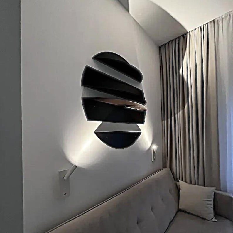May include: A modern wall-mounted light fixture with a unique design. The fixture is composed of several black, curved, layered elements, creating a three-dimensional effect. The fixture is mounted on a white wall, with a soft light illuminating the area below.