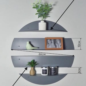 May include: Three gray, semi-circular wall shelves, each 66cm wide, are mounted on a light gray wall. The top shelf holds a white vase with greenery. The middle shelf displays a framed photo and a green bird figurine. The bottom shelf has two candles and a small vase.