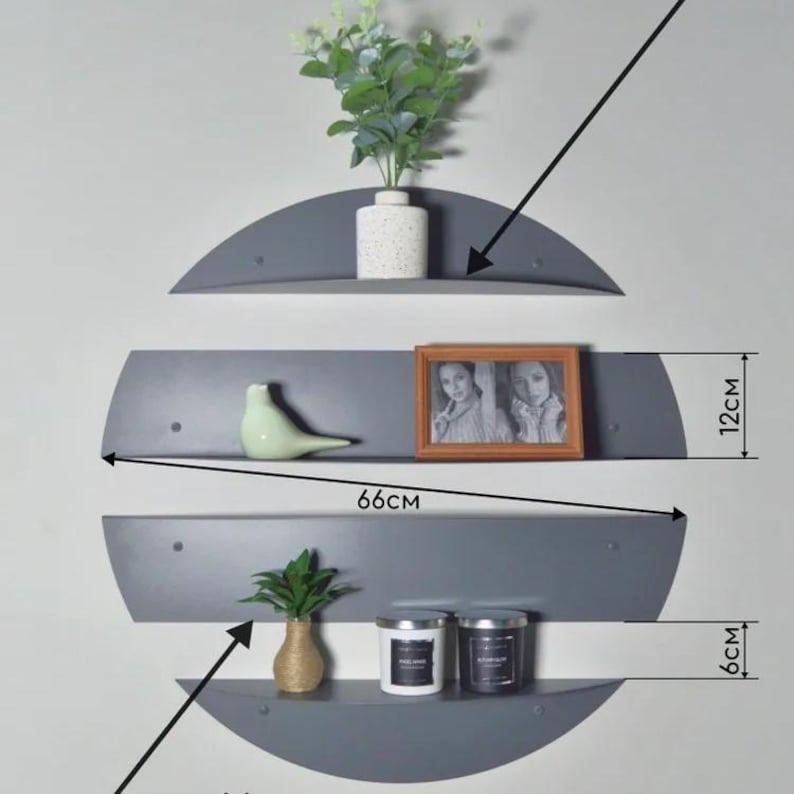 May include: Three gray, semi-circular wall shelves, each 66cm wide, are mounted on a light gray wall. The top shelf holds a white vase with greenery. The middle shelf displays a framed photo and a green bird figurine. The bottom shelf has two candles and a small vase.