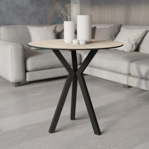 May include: A round side table with a light wood grain top and black metal legs. The table is 76 cm in diameter and features a unique X-shaped leg design. Two white vases and two small white bowls are on the table.
