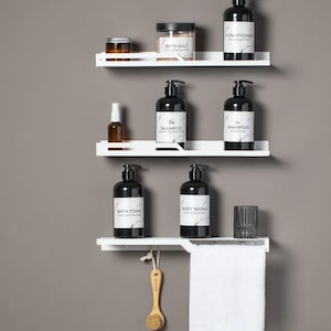 Puede incluir: Tres estantes blancos con detalles metálicos negros montados en una pared gris. Los estantes están llenos de varios productos de baño, incluyendo botellas de champú, acondicionador, gel de baño, espuma de baño y sales de baño. Una toalla blanca cuelga de un gancho debajo de los estantes.