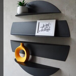 May include: A set of four dark gray metal wall shelves in a unique, semi-circular design. The shelves hold a small potted plant, a framed picture with text, and a yellow vase. The shelves are mounted on a light gray wall.